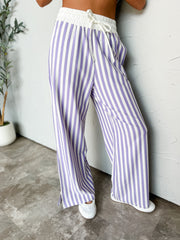 A person wearing loose, wide-leg striped pants with vertical purple and white stripes, a white elastic drawstring waist, and white slip-on shoes, standing against a light wall.