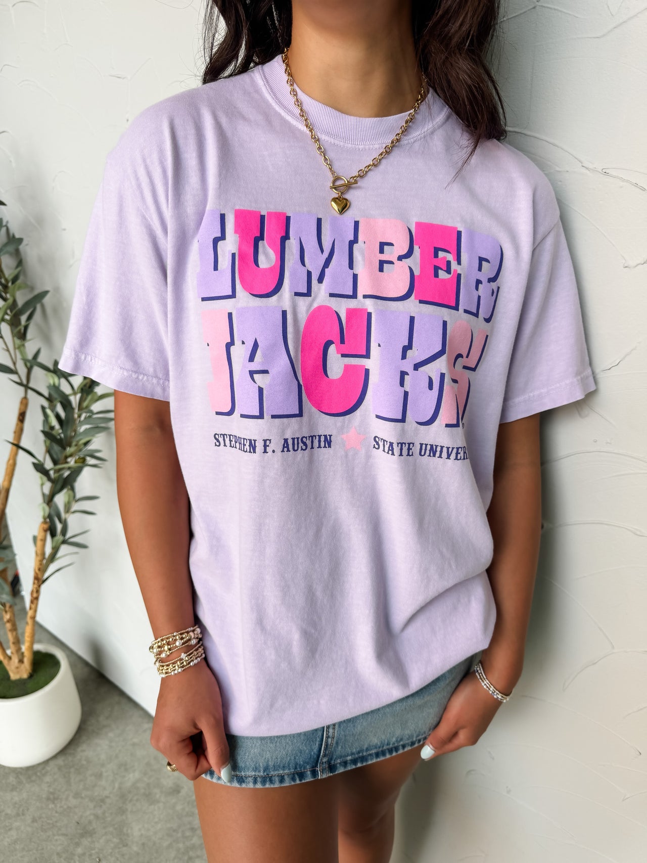 Person wearing a light purple t-shirt with colorful text, standing against a white wall.