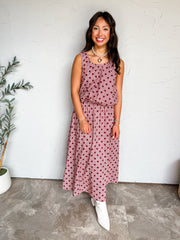 A full-length photograph of a smiling woman with long dark wavy hair holding a strand of her hair while posing against a textured white wall next to a potted olive tree. She is wearing a two-piece matching set made from 100% cotton, featuring a burgundy red and white micro-gingham check print overlaid with large dark red polka dots, including a sleeveless scoop-neck tank top with an elasticated hem and a matching high-waisted A-line maxi skirt, styled with white pointed-toe ankle boots, a pearl choker neckl