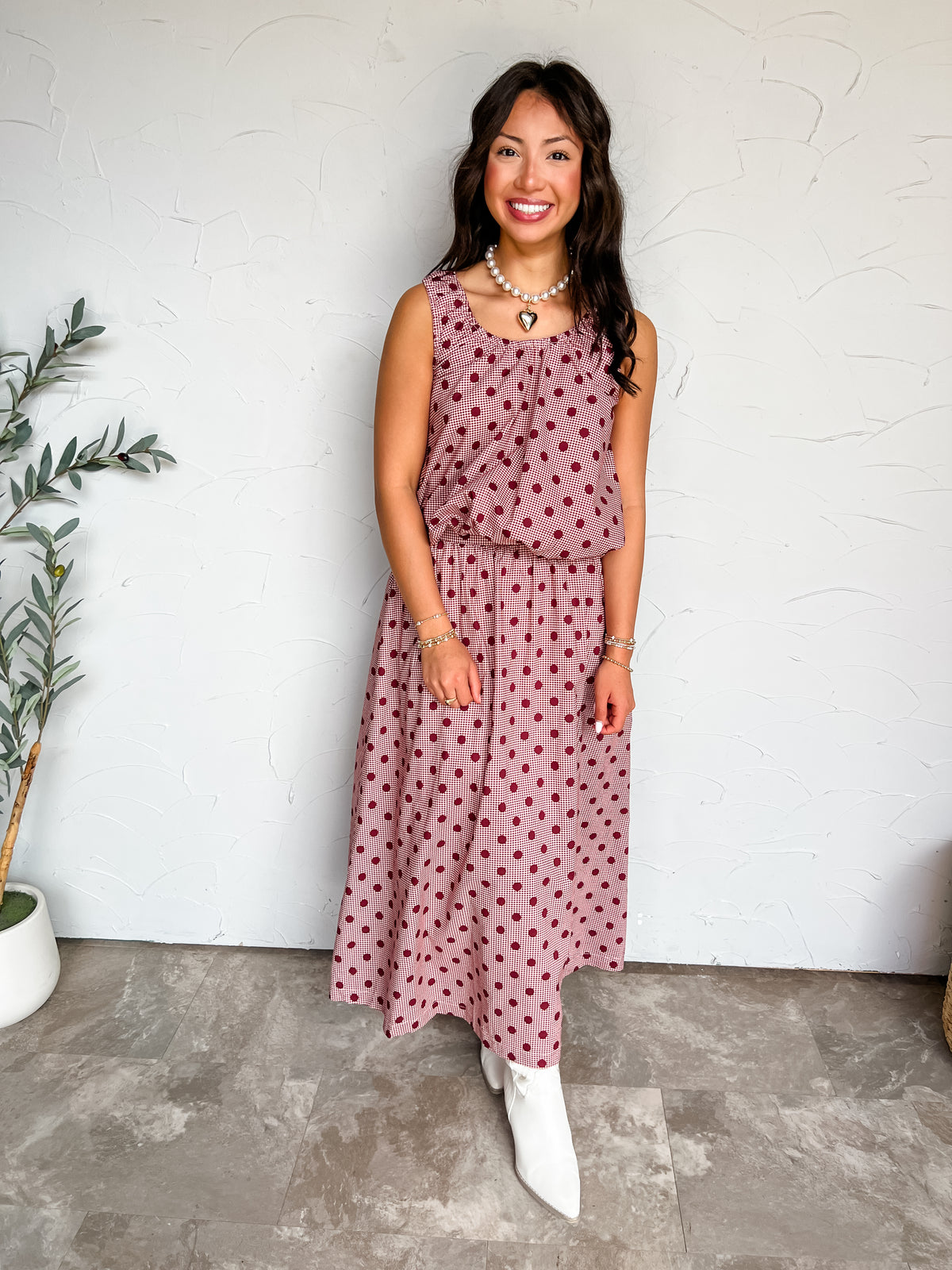 A full-length photograph of a smiling woman with long dark wavy hair holding a strand of her hair while posing against a textured white wall next to a potted olive tree. She is wearing a two-piece matching set made from 100% cotton, featuring a burgundy red and white micro-gingham check print overlaid with large dark red polka dots, including a sleeveless scoop-neck tank top with an elasticated hem and a matching high-waisted A-line maxi skirt, styled with white pointed-toe ankle boots, a pearl choker neckl