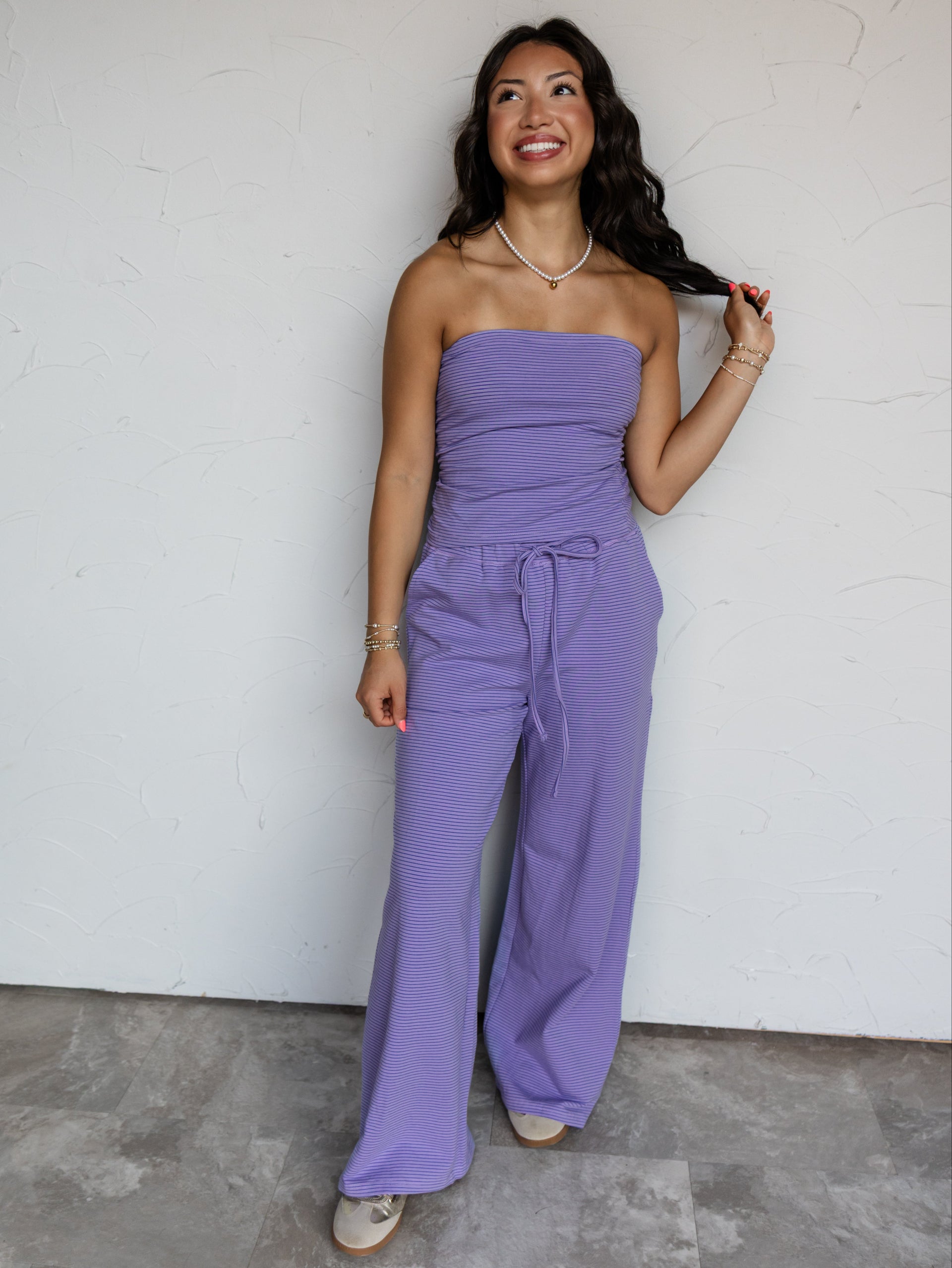 A full-length photograph of a smiling woman with long wavy dark brown hair posing with one hand on her hip against a textured white wall. She is wearing a lavender purple micro-striped two-piece knit set made from a 95% cotton and 5% spandex blend fabric, featuring a strapless tube top with side ruching and matching drawstring wide-leg pants, styled with neutral sneakers and a pearl choker necklace.