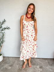 A full-length photograph of a smiling young woman with long wavy dark blonde hair posing against a textured white wall next to a potted olive tree. She is wearing a sleeveless cream midi dress with a red floral pattern made from a 97% polyester and 3% spandex blend, featuring wide shoulder straps, a gathered ruched bust, a fitted waist, and a multi-tiered ruffle skirt, styled with clear slide heels, a chunky red beaded necklace, a delicate silver cross necklace, and a simple beaded bracelet.