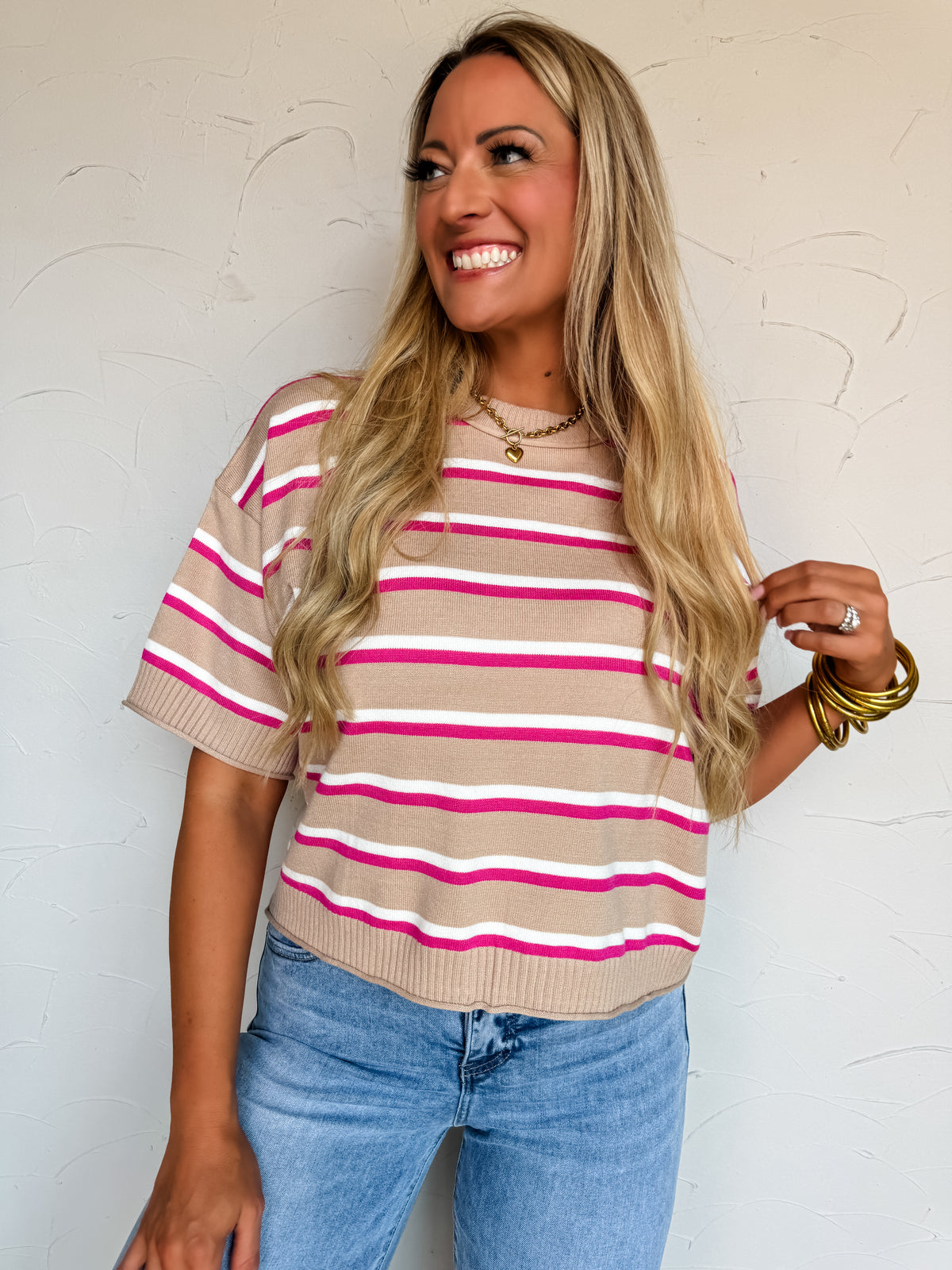 A waist-up photograph of a smiling blonde woman looking off to the side while holding a strand of her hair against a textured white wall. She is wearing a short-sleeved fine-knit sweater made from a 45% viscose, 28% polyester, and 27% nylon blend, featuring horizontal stripes in hot pink, white, and tan, styled with light-wash jeans, stacked gold bangles, a gold chain heart pendant necklace, and a ring.