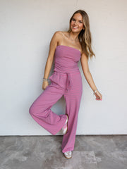 A full-length photograph of a smiling woman with long blonde hair posing playfully with one leg raised against a textured white wall. She is wearing a pink and blue micro-striped two-piece knit set made from a 95% cotton and 5% spandex blend fabric, featuring a strapless tube top with side ruching and matching drawstring wide-leg pants, styled with pink-striped sneakers and silver jewelry.