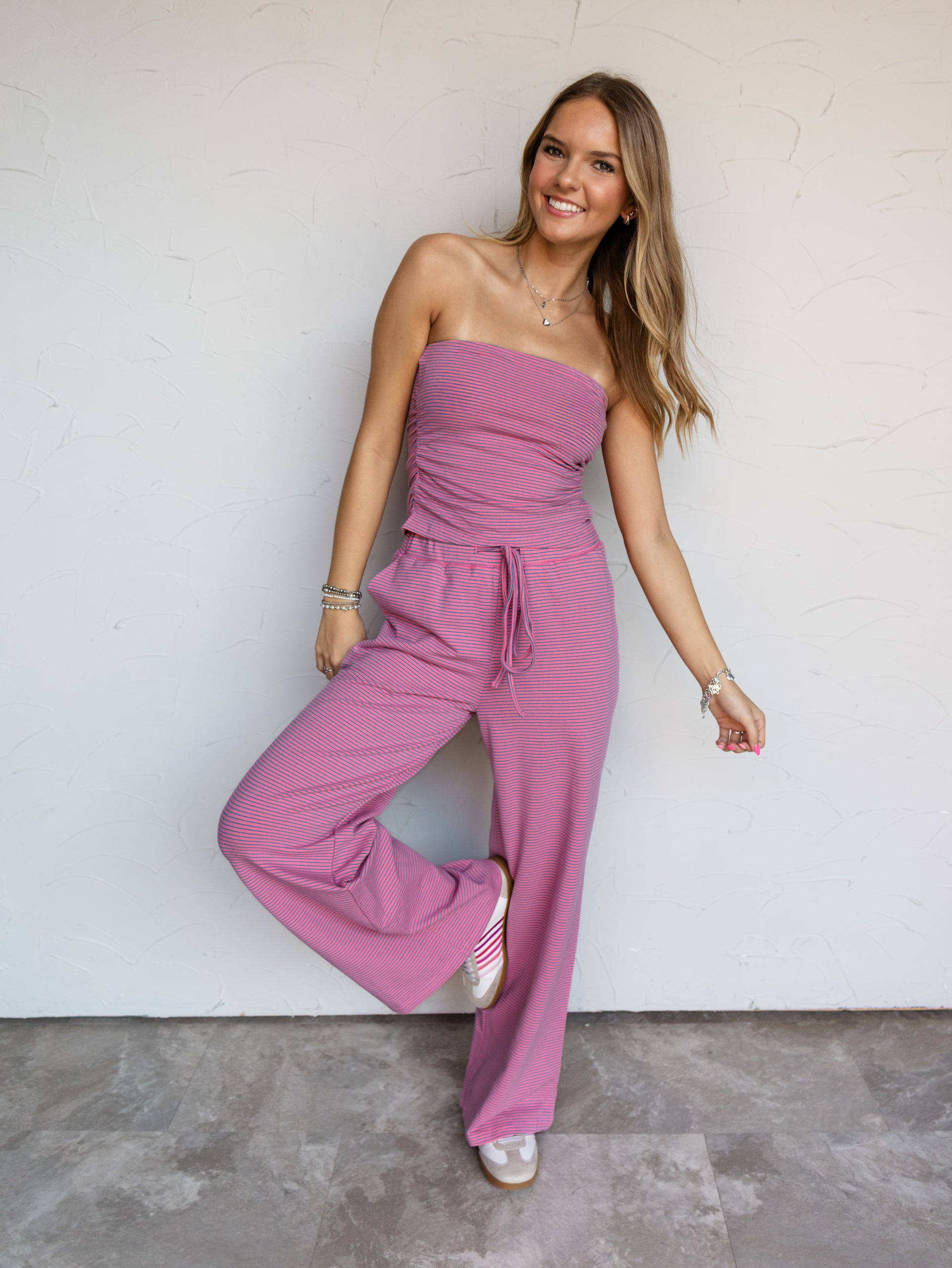 A full-length photograph of a smiling woman with long blonde hair posing playfully with one leg raised against a textured white wall. She is wearing a pink and blue micro-striped two-piece knit set made from a 95% cotton and 5% spandex blend fabric, featuring a strapless tube top with side ruching and matching drawstring wide-leg pants, styled with pink-striped sneakers and silver jewelry.