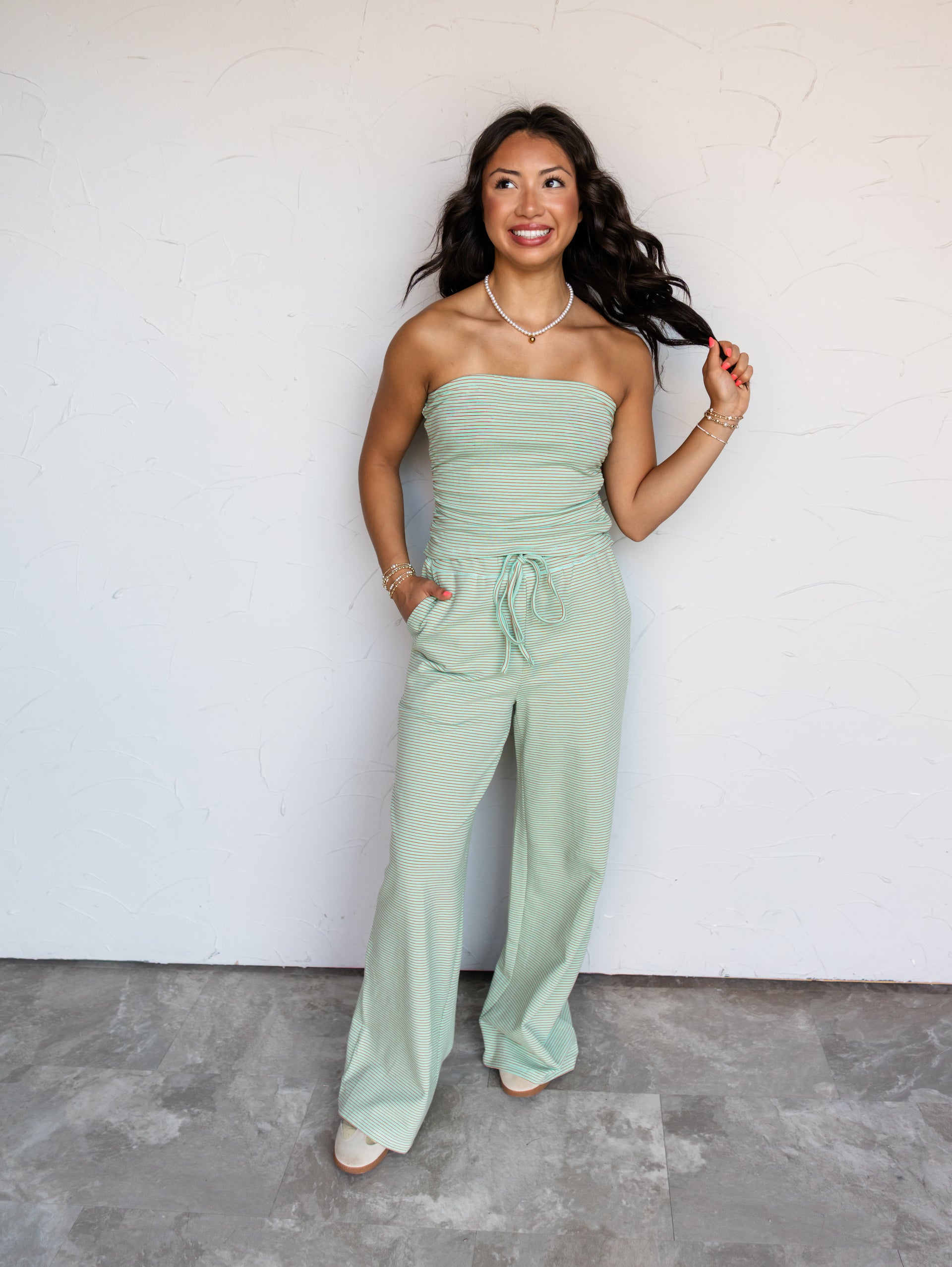 A full-length photograph of a smiling woman with long wavy dark brown hair holding a strand of her hair while posing against a textured white wall. She is wearing a mint green and orange micro-striped two-piece knit set made from a 95% cotton and 5% spandex blend fabric, featuring a strapless tube top with side ruching and matching drawstring wide-leg pants with side pockets, styled with neutral sneakers and a pearl choker necklace.