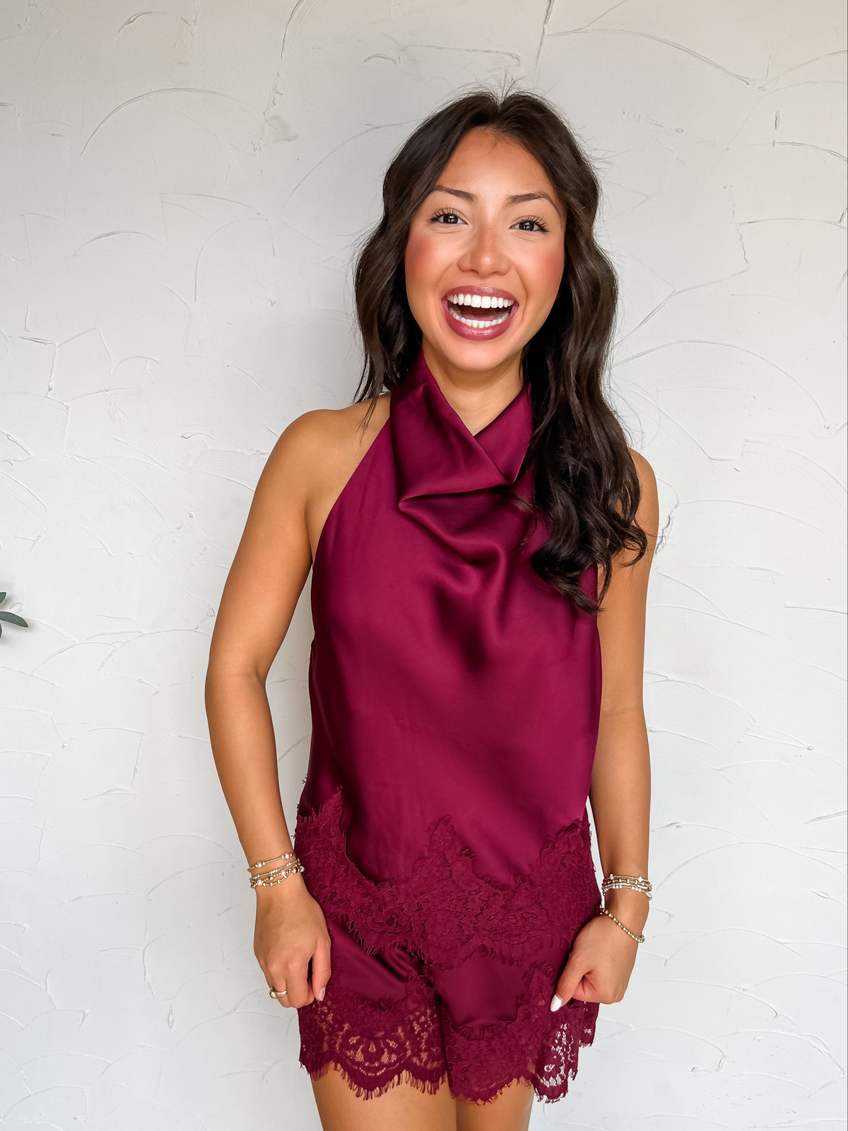 A three-quarter length photograph of a smiling woman with long dark wavy hair posing joyfully against a textured white wall. She is wearing a two-piece burgundy satin matching set made from 100% polyester, featuring a sleeveless high-neck draped cowl halter top accented with floral eyelash lace trim at the hem, paired with matching satin shorts with a wide scalloped eyelash lace hemline, styled with white cowboy boots, gold rings, and delicate beaded bracelets.