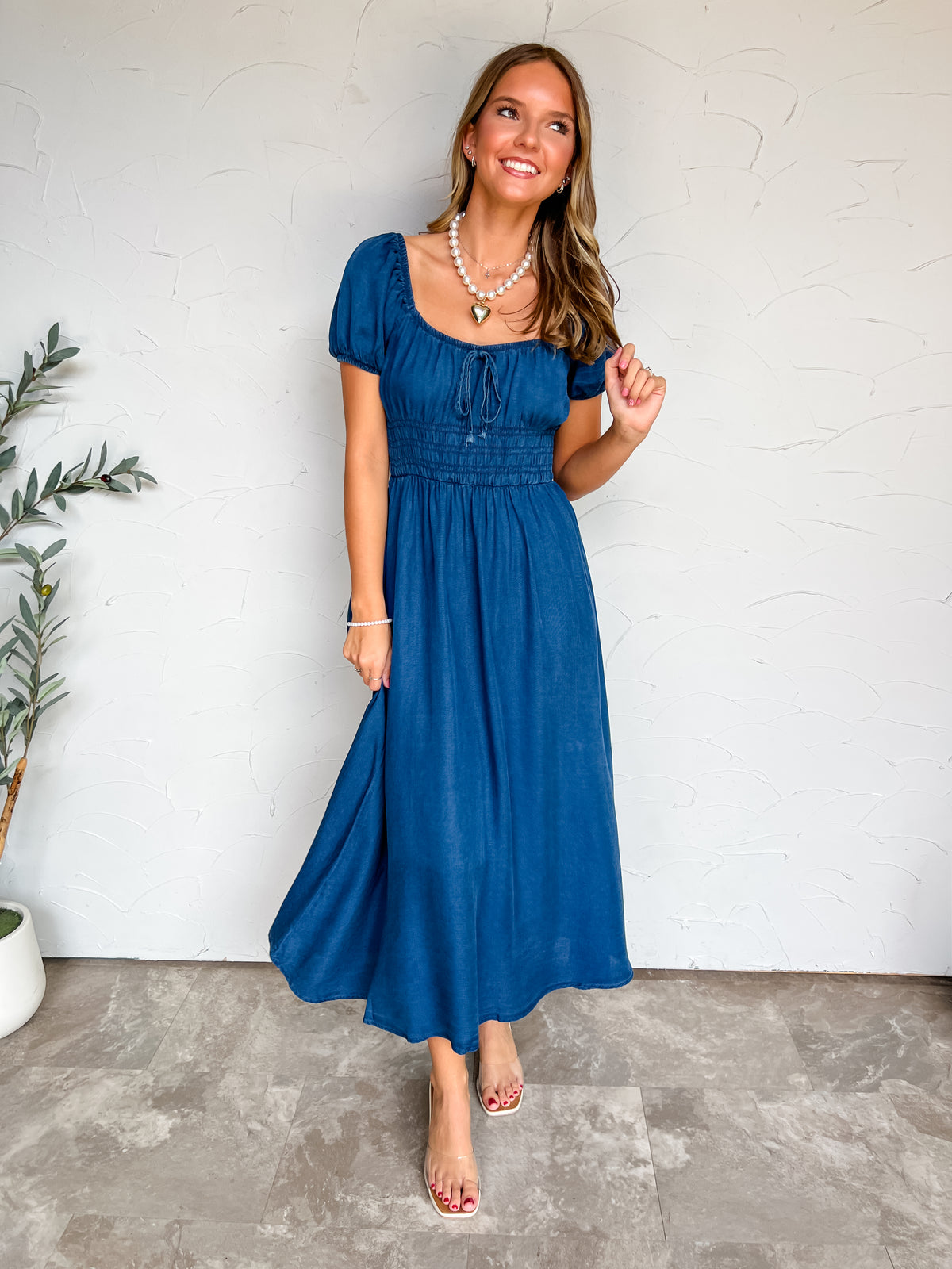 A full-length photograph of a smiling young woman with long wavy dark blonde hair twirling her dress slightly while posing against a textured white wall next to a potted olive tree. She is wearing a dark denim blue midi dress made from 100% lyocell, featuring a scoop neckline with a small center bust tie detail, short elasticized puff sleeves, a wide smocked elastic waist, and a flared A-line skirt, styled with clear double-strap slide sandals, a pearl necklace featuring a gold heart pendant, a silver cross