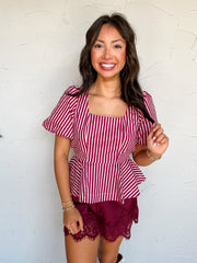 A three-quarter length photograph of a smiling woman with long dark wavy hair twirling a section of her hair while posing against a textured white wall. She is wearing a short-sleeved cotton blouse featuring vertical burgundy and white stripes made from 100% cotton, with a square neckline, short puff sleeves, and a flared peplum hem, paired with burgundy satin shorts with eyelash lace trim, styled with white cowboy boots, gold rings, and stacked beaded bracelets.