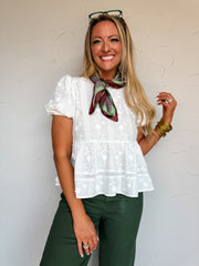 A waist-up photograph of a smiling blonde woman with green glasses resting on her head, posing against a textured white wall. She is wearing a short-sleeved white peplum top made from 100% cotton, featuring floral eyelet embroidery, crochet lace inserts, short puff sleeves, and a tiered peplum hem, accessorized with a brown and green printed neck scarf tied around the collar, dark green pants, stacked gold bangles, and rings.