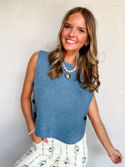 A three-quarter length photograph of a smiling young woman with long wavy brown hair with blonde highlights posing with one hand in her pocket against a textured white wall. She is wearing a sleeveless dusty blue fine-knit sweater vest made from a 58% polyester, 18% acrylic, 15% nylon, 6% wool, and 2% spandex blend, featuring a crew neckline, boxy fit, and side slits held together by dark tortoiseshell buttons, paired with cream high-waisted pants with blue floral stripe printing, styled with a pearl neckla