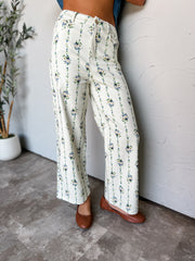A photograph from the waist down of a model leaning against a textured off-white wall in a studio setting next to a potted olive tree. She is wearing high-waisted, wide-leg cream pants adorned with vertical blue and green floral vine stripes made from a 97% cotton and 3% spandex blend, featuring belt loops, a front zip fly with button closure, and a wide-leg cut, styled with brown leather ballet flats and a blue top.