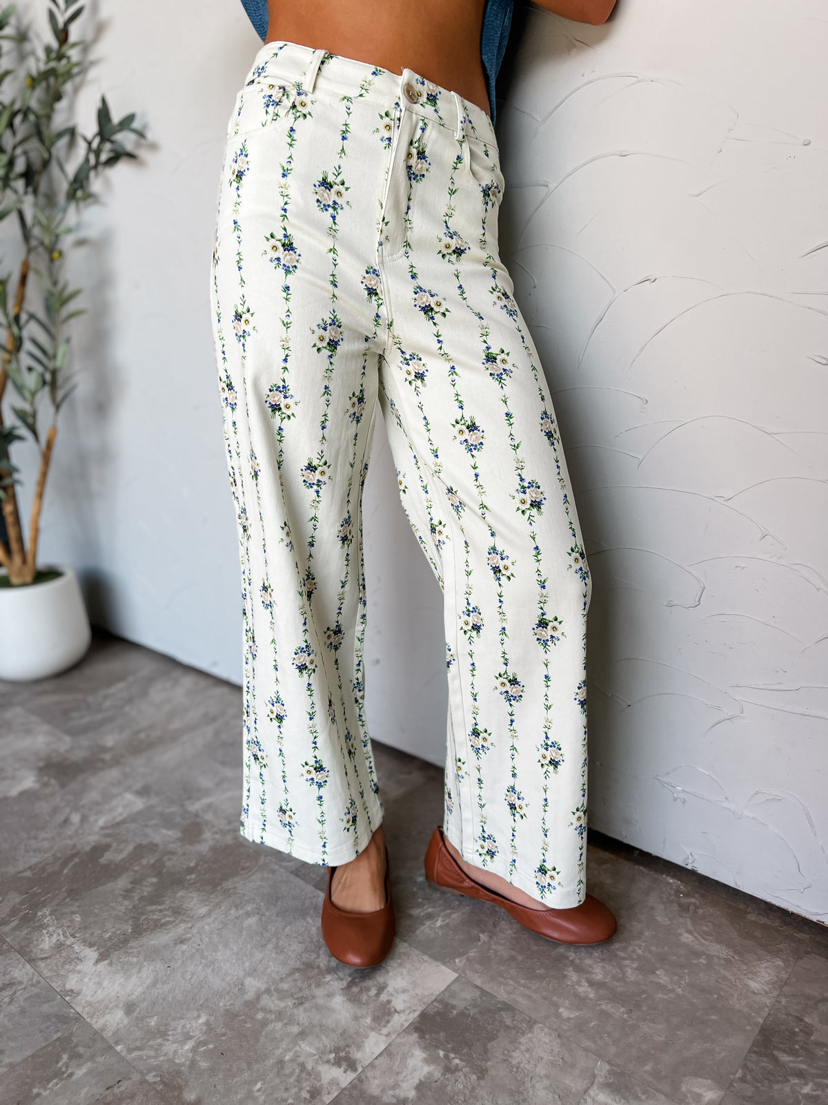 A photograph from the waist down of a model leaning against a textured off-white wall in a studio setting next to a potted olive tree. She is wearing high-waisted, wide-leg cream pants adorned with vertical blue and green floral vine stripes made from a 97% cotton and 3% spandex blend, featuring belt loops, a front zip fly with button closure, and a wide-leg cut, styled with brown leather ballet flats and a blue top.