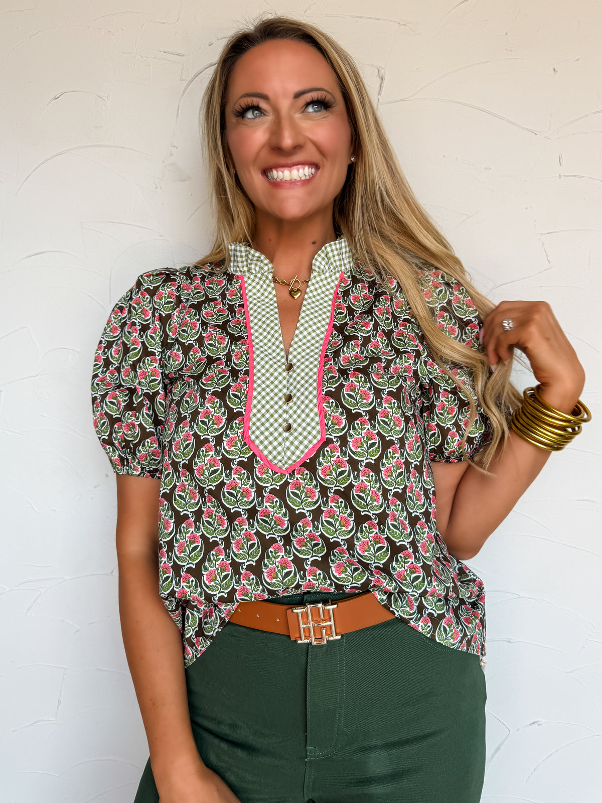 A waist-up photograph of a smiling blonde woman looking upward while touching a strand of her hair against a textured white wall. She is wearing a short-sleeved cotton blouse featuring a brown and olive green background with a pink floral print, a contrasting green and white gingham split-neck placket trimmed with hot pink piping, a ruffle collar, and puff sleeves, tucked into forest green pants with a tan leather belt and styled with stacked gold bangles and a gold heart pendant necklace.