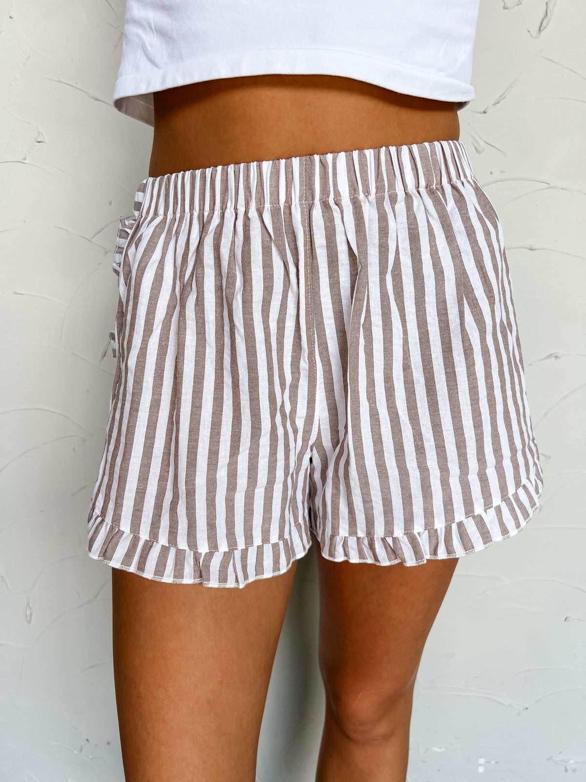 A close-up photograph from the waist to the mid-thigh of a model posing against a textured off-white wall. She is wearing high-waisted taupe brown and white vertical striped shorts made from 100% cotton, featuring an elastic gathers waistband, a ruffle-trimmed hem, and side pocket detail, paired with a fitted white crop top.
