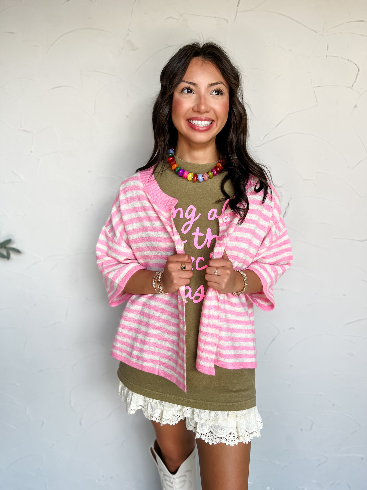 A three-quarter length photograph of a smiling woman with long dark wavy hair looking away while holding open the lapels of her sweater against a textured white wall. She is wearing a short-sleeved button-front knit cardigan featuring horizontal bright pink and cream stripes made from a 76% polyester, 15% acrylic, 6% wool, and 3% spandex blend, layered over an olive green graphic tee with pink lettering and a cream lace-trimmed skirt, styled with a colorful beaded choker necklace and stacked bracelets.