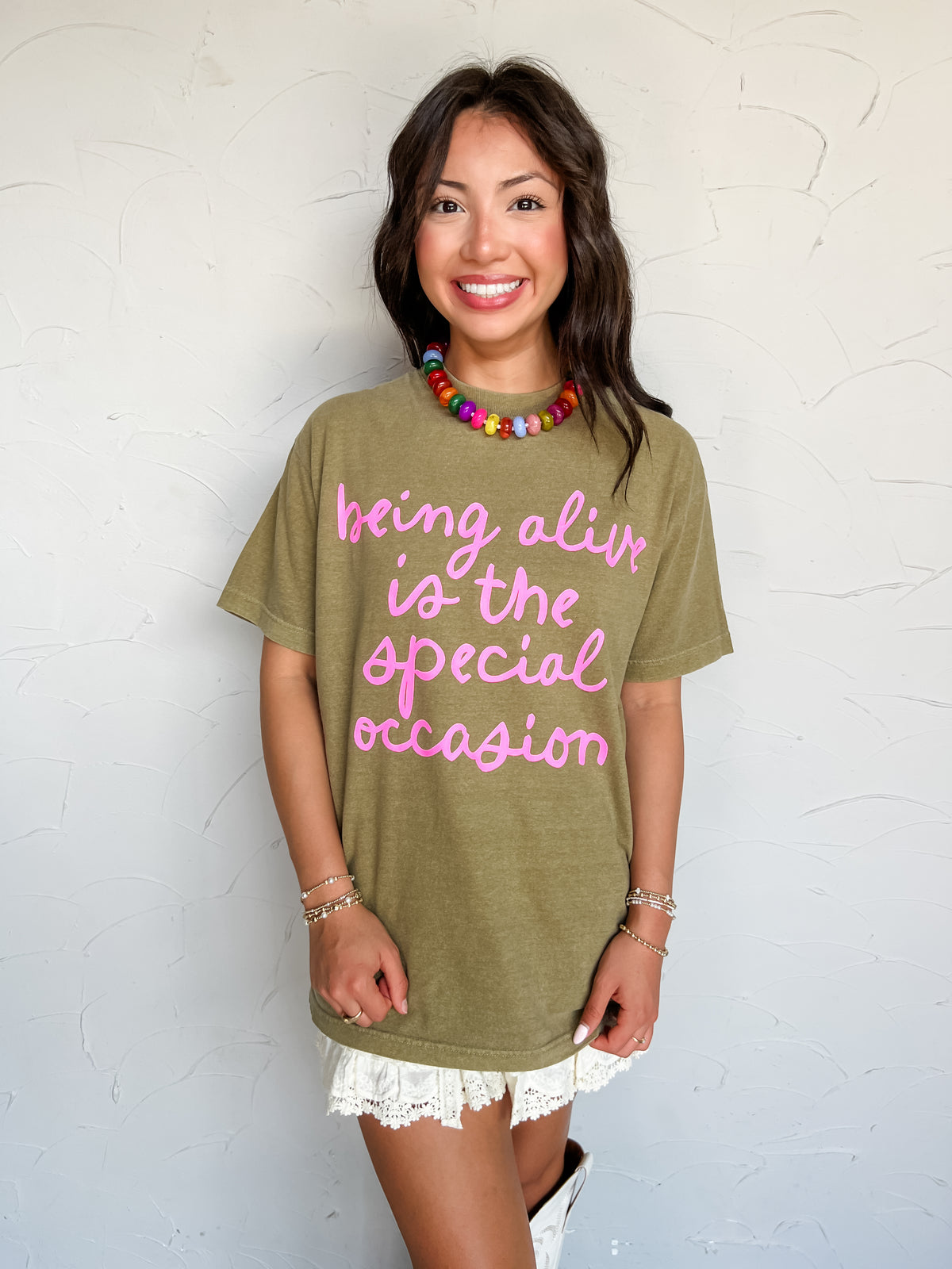 A woman wearing an oversized olive green graphic t-shirt with pink text that reads 'being alive is the special occasion'. She is standing against a textured white wall.