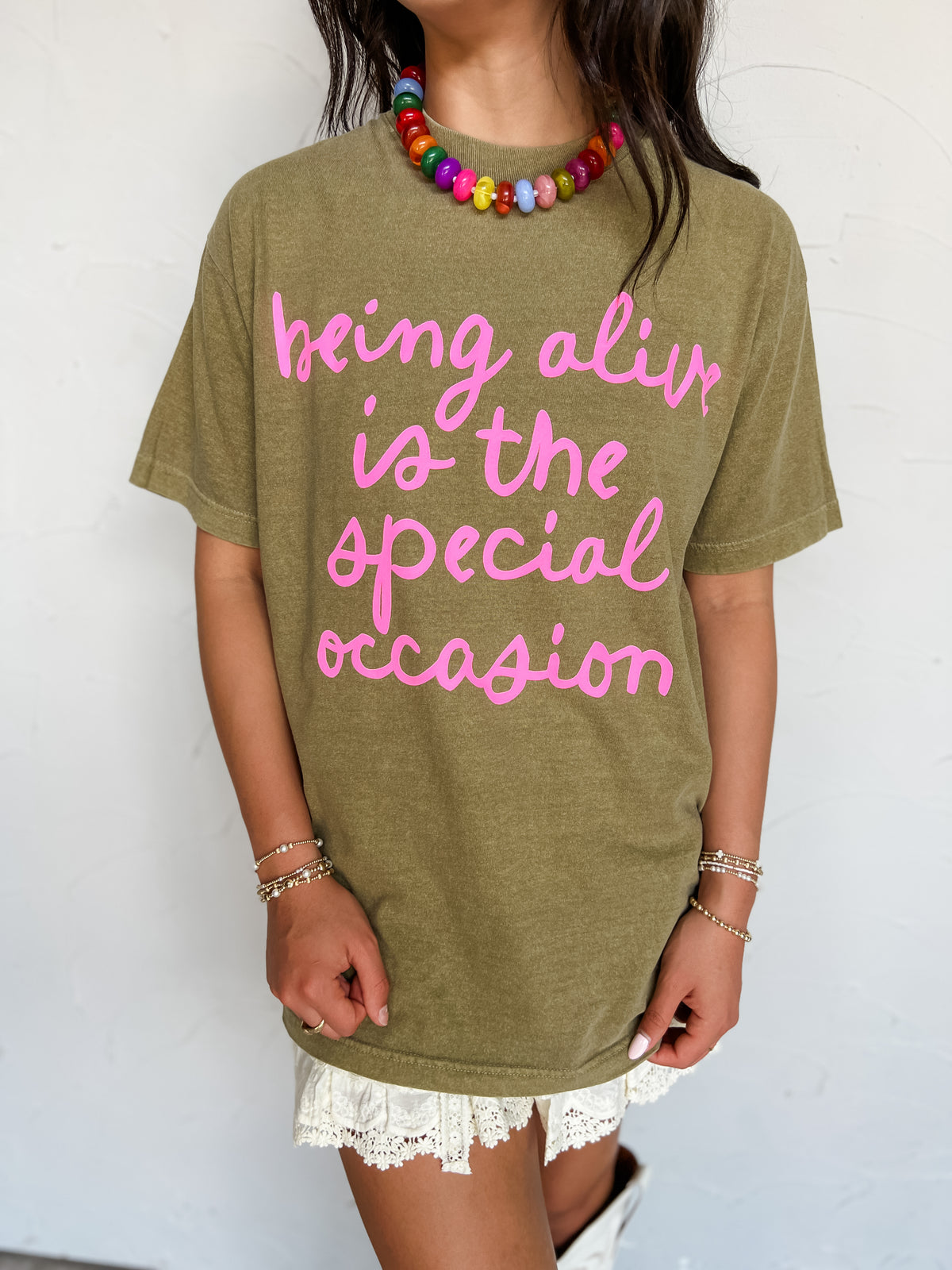 A woman wearing an olive green graphic t-shirt with pink text that reads 'being alive is the special occasion'. She is also wearing a colorful beaded necklace and white lace shorts, standing against a white wall.
