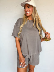 A waist-up photograph of a smiling blonde woman looking off to the side while holding a lock of her long hair against a textured white wall. She is wearing a matching two-piece mineral-washed charcoal taupe lounge set made from 100% cotton, featuring an oversized short-sleeved crewneck tee and relaxed pull-on shorts, accessorized with a beige trucker hat that reads "Praise Him anyway.", stacked gold bangles, a gold heart pendant chain necklace, and various rings.