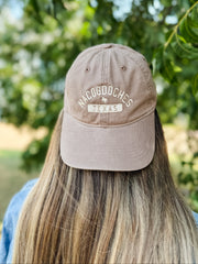 A person viewed from behind wearing a dusty taupe baseball cap with 'NACOGDOCHES TEXAS' embroidered on the front, set against a soft-focus background of green trees.