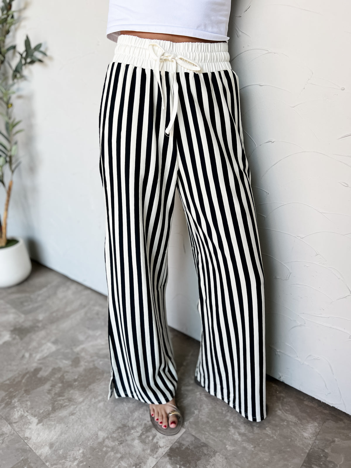 A full-length photograph from the waist down of a model leaning against a textured white wall in a studio setting next to a potted olive tree. She is wearing high-waisted wide-leg pants featuring bold vertical black and off-white stripes, made from a 95% cotton and 5% spandex blend, with a smocked cream elastic waistband with an adjustable drawstring and subtle side slits at the hem, paired with a white top and clear strap slide sandals.