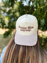 A person viewed from behind wearing a cream-colored trucker-style cap with brown embroidered text reading 'Praise Him anyway.' and a brown-and-white gingham patterned visor, set against a blurred outdoor background of green foliage.