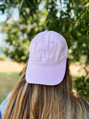 A lavender purple baseball cap with 'SEASU' and a Texas longhorn logo embroidered on the front, worn by a person with long brown hair outdoors with a green, blurred background.