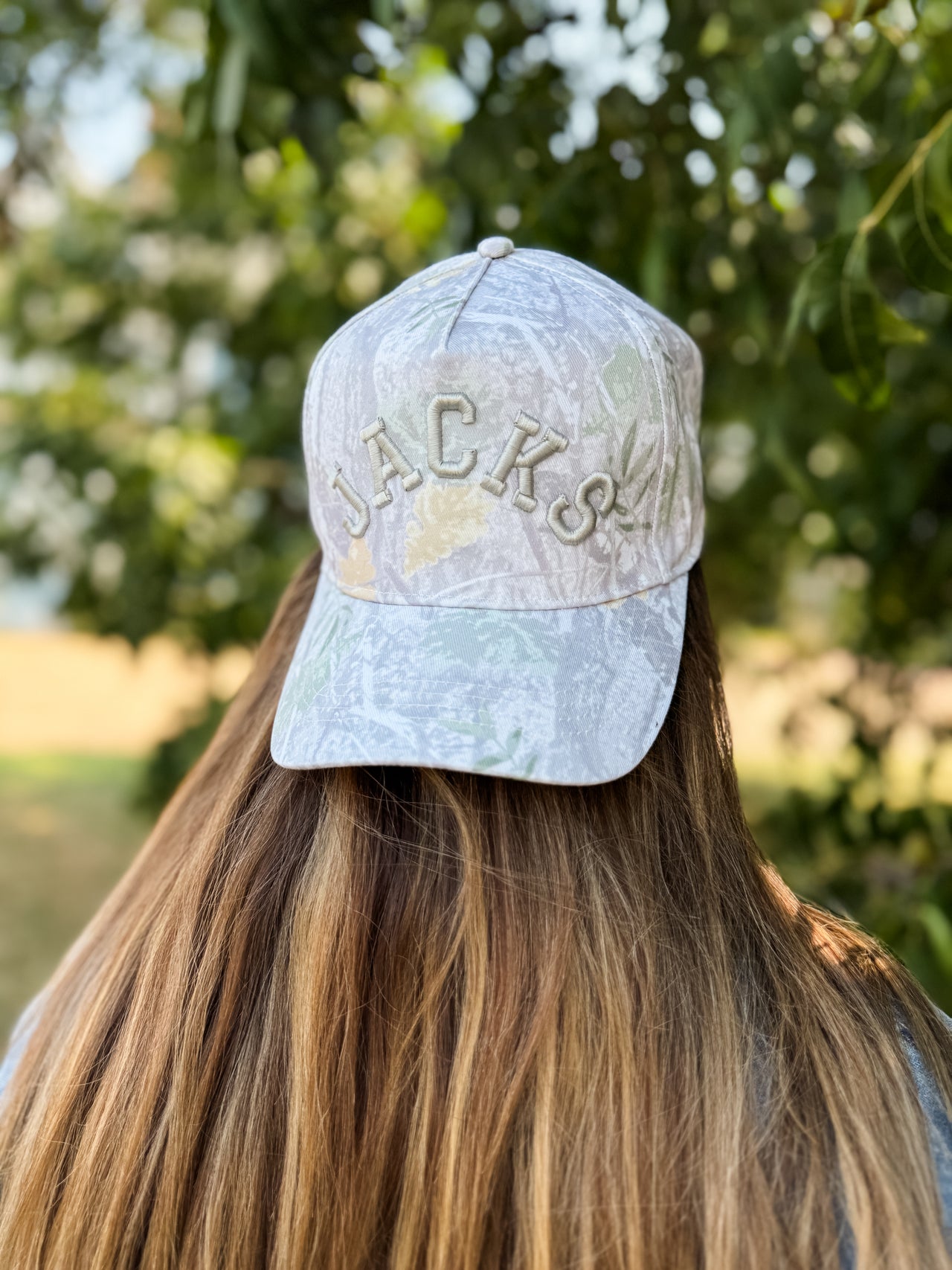 A person viewed from behind wearing a light gray camouflage baseball cap with 'JACKS' embroidered on the front, set against a blurred outdoor background of green trees.