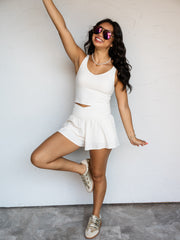 A full-length photograph of a smiling woman with long dark wavy hair raising one arm and posing playfully on one leg against a textured white wall. She is wearing an ivory two-piece activewear set made from an 84% polyester and 16% elastane blend fabric, featuring a sleeveless V-neck crop tank top and matching high-waisted shorts with a crossover waistband, styled with metallic sneakers, aviator sunglasses, and a pearl choker necklace.