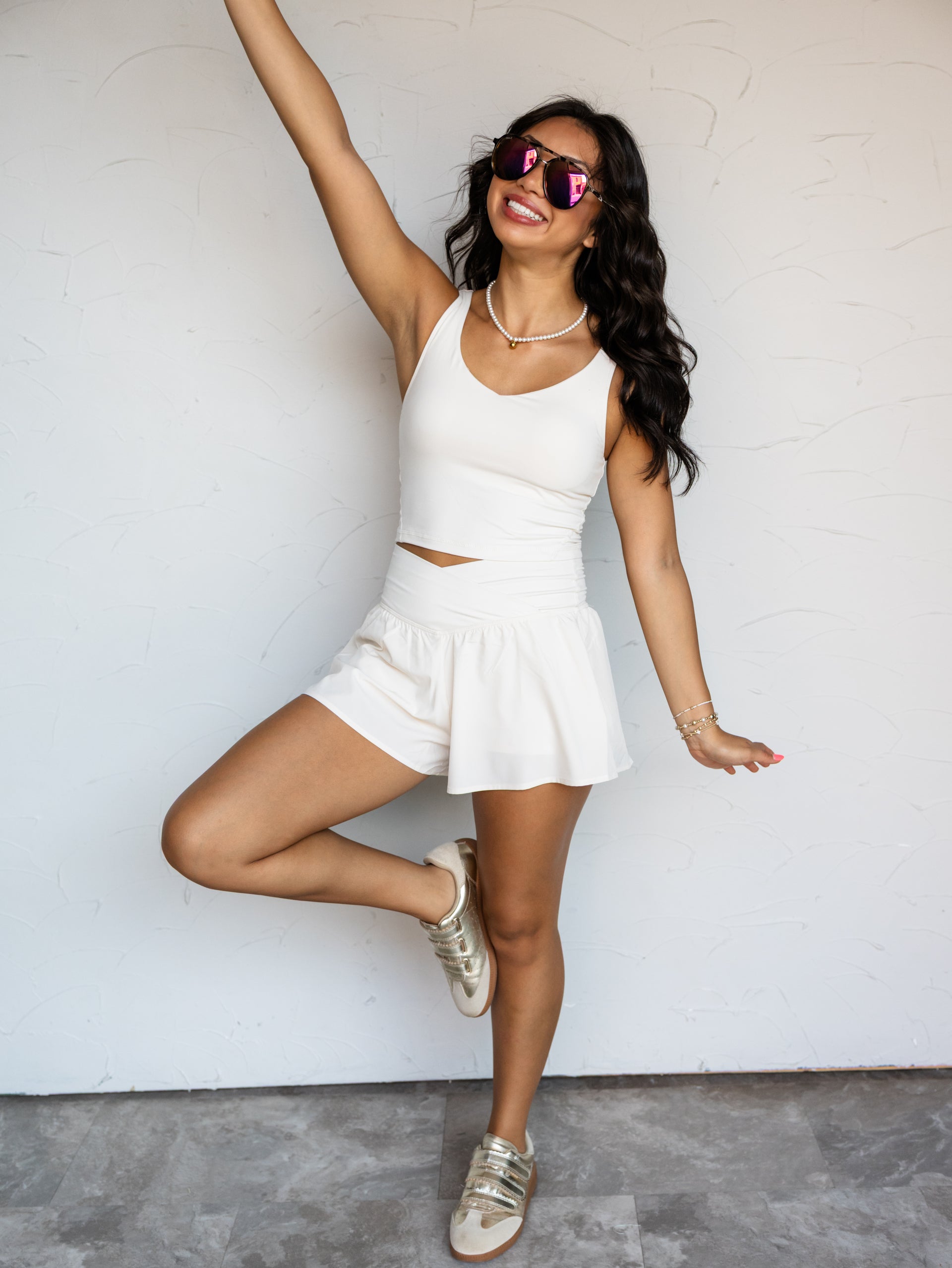 A full-length photograph of a smiling woman with long dark wavy hair raising one arm and posing playfully on one leg against a textured white wall. She is wearing an ivory two-piece activewear set made from an 84% polyester and 16% elastane blend fabric, featuring a sleeveless V-neck crop tank top and matching high-waisted shorts with a crossover waistband, styled with metallic sneakers, aviator sunglasses, and a pearl choker necklace.