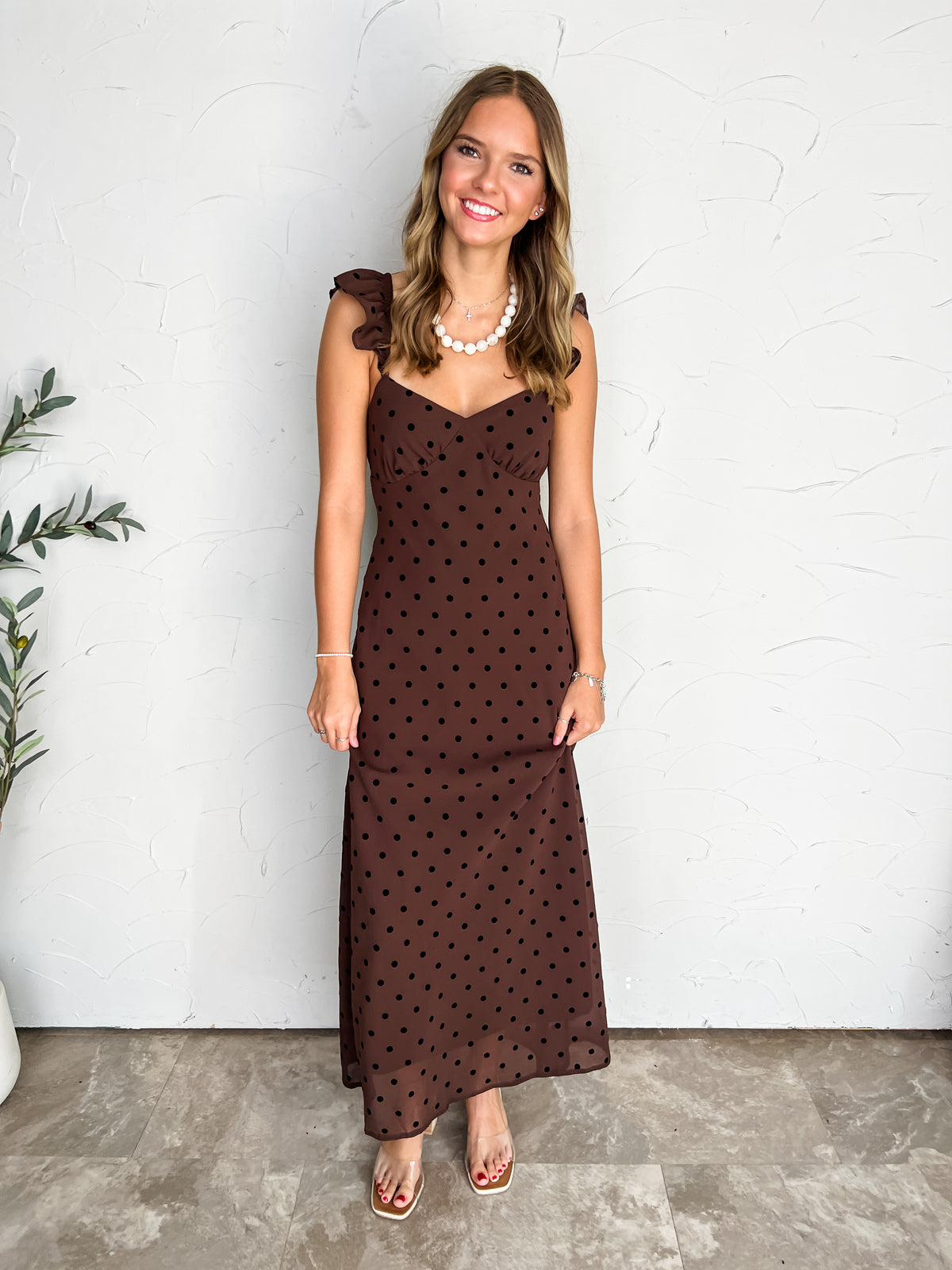 A full-length photograph of a smiling young woman with light brown wavy hair posing against a textured off-white wall next to a potted olive plant. She is wearing a sleeveless chocolate brown maxi dress made from 100% polyester in a black polka dot pattern, featuring ruffled shoulder straps, a structured sweetheart neckline, and a flowing skirt hem, styled with clear slide heels, a white beaded pearl necklace with a small cross pendant, delicate silver bracelets, and stud earrings