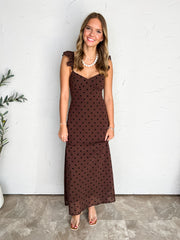 A full-length photograph of a smiling young woman with light brown wavy hair posing against a textured off-white wall next to a potted olive plant. She is wearing a sleeveless chocolate brown maxi dress made from 100% polyester in a black polka dot pattern, featuring ruffled shoulder straps, a structured sweetheart neckline, and a flowing skirt hem, styled with clear slide heels, a white beaded pearl necklace with a small cross pendant, delicate silver bracelets, and stud earrings
