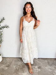 A full-length photograph of a smiling brunette woman holding a strand of her long wavy hair while posing against a textured off-white wall next to a potted olive plant. She is wearing an off-white sleeveless midi dress made from 100% polyester, featuring a floral lace overlay, sweetheart neckline, delicate lace straps, and a tiered skirt, styled with gold metallic slide sandals, a round gold pendant necklace, stacked gold bracelets, and rings.