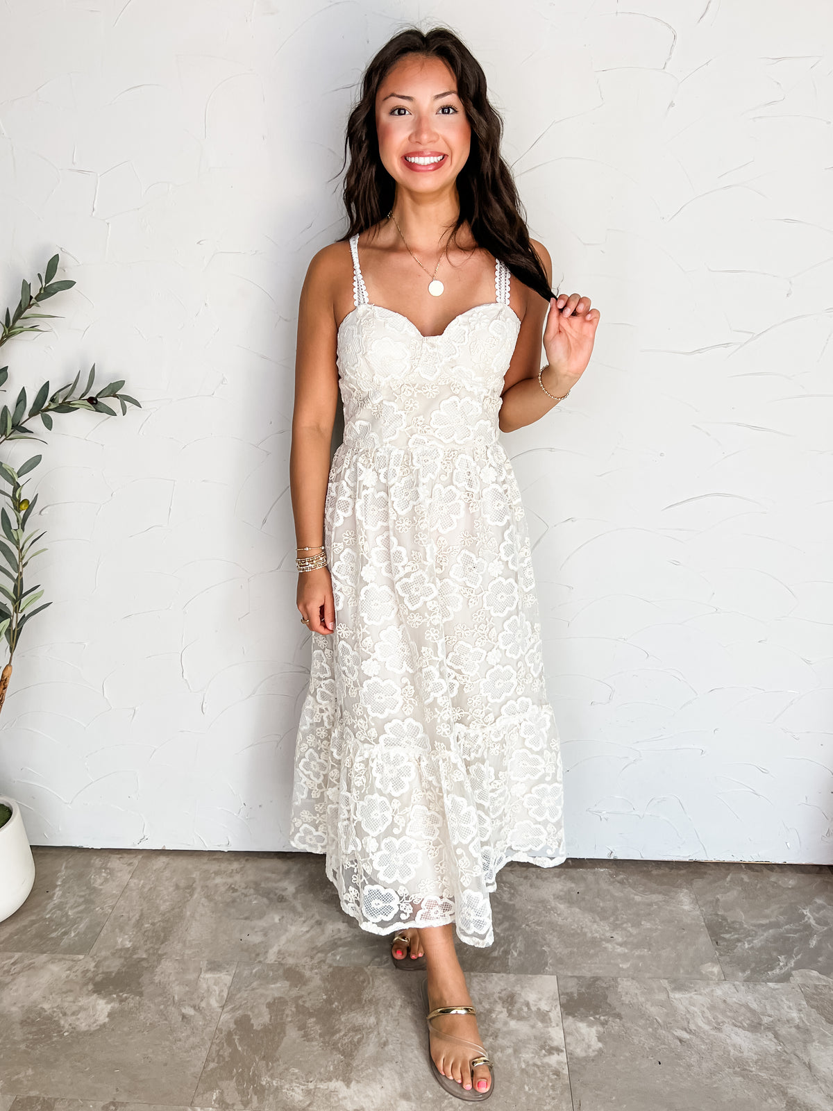 A full-length photograph of a smiling brunette woman holding a strand of her long wavy hair while posing against a textured off-white wall next to a potted olive plant. She is wearing an off-white sleeveless midi dress made from 100% polyester, featuring a floral lace overlay, sweetheart neckline, delicate lace straps, and a tiered skirt, styled with gold metallic slide sandals, a round gold pendant necklace, stacked gold bracelets, and rings.