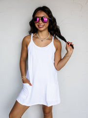A three-quarter length photograph of a smiling woman with long dark wavy hair holding a strand of her hair while posing with one hand in her pocket against a textured white wall. She is wearing a crisp white sleeveless mini dress made from a 95% cotton and 5% spandex blend fabric, featuring a scoop neckline, thin shoulder straps, and two low front patch pockets, styled with aviator sunglasses, a pearl choker necklace, and stacked bracelets.