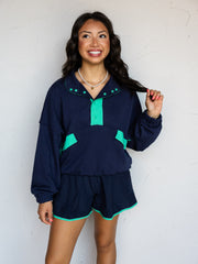 A three-quarter length photograph of a smiling woman with long wavy dark brown hair holding a strand of her hair while posing against a textured white wall. She is wearing a dark navy blue long-sleeved sweatshirt with bright mint green contrast trim on the snap placket, collar, and front flap pockets, made from a 60% cotton and 40% polyester blend fabric, styled with matching navy shorts with mint trim and a pearl choker necklace.