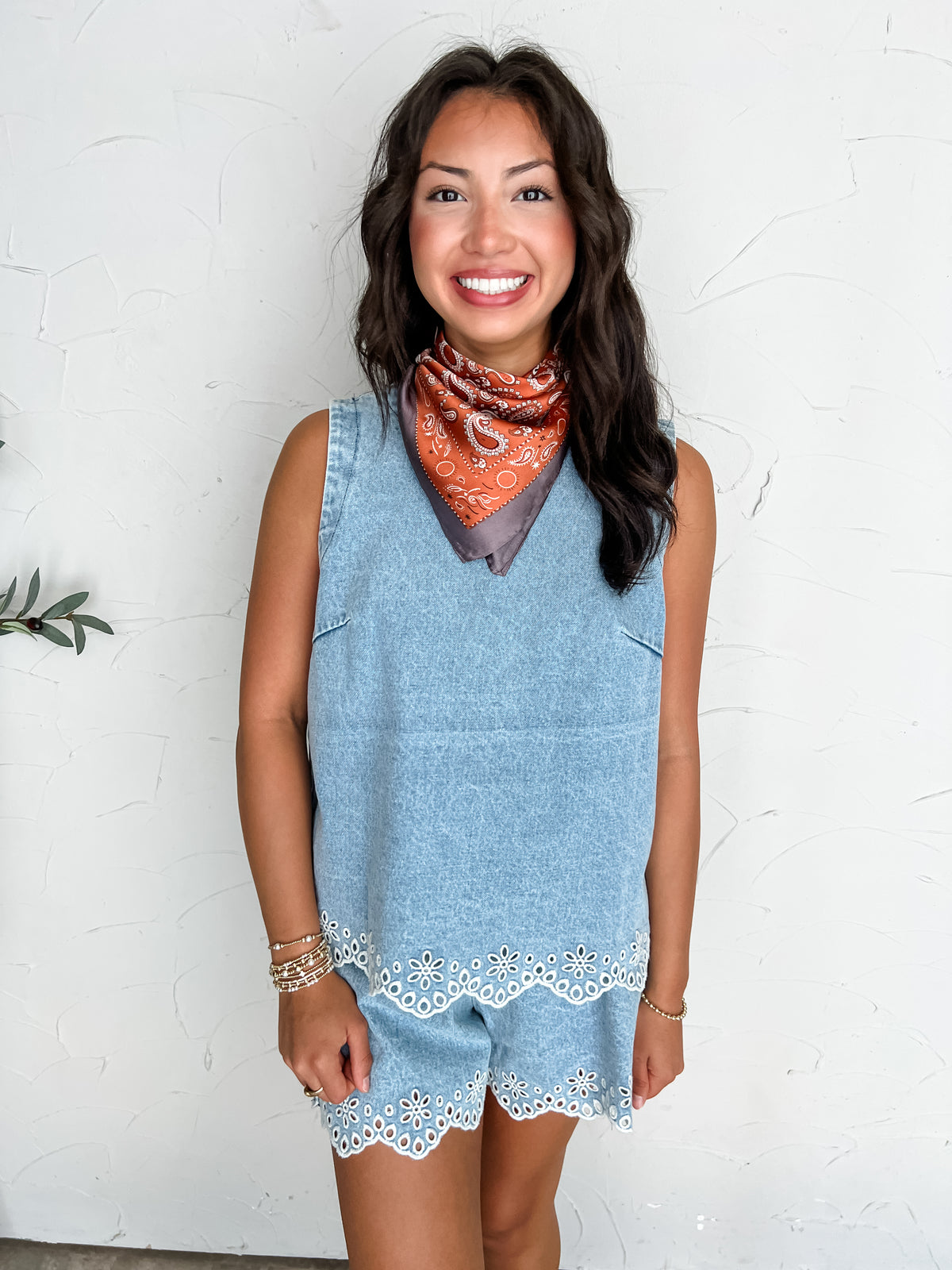 A portrait photograph of a smiling brunette woman posing against a textured off-white wall. She is wearing a light wash denim two-piece matching set made from 100% cotton, featuring a sleeveless crewneck tunic top decorated with white embroidered floral eyelet scalloped trim along the hem, paired with matching denim shorts with identical eyelet trim along the leg openings, styled with a terracotta orange paisley neck scarf, stacked gold beaded bracelets, and gold rings.