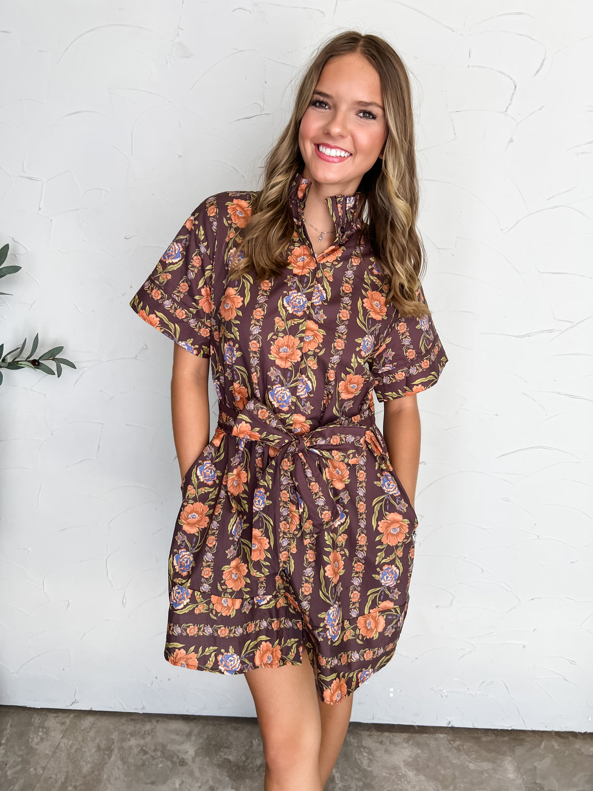 A portrait photograph of a smiling young woman with light brown wavy hair posing with her hands in her pockets against a textured off-white wall. She is wearing a short-sleeved romper made from 100% cotton featuring a chocolate brown background with an orange and blue floral pattern, a high stand-up collar, cuffed sleeves, and a matching self-tie waist belt, styled with a delicate silver cross pendant necklace and stud earrings.