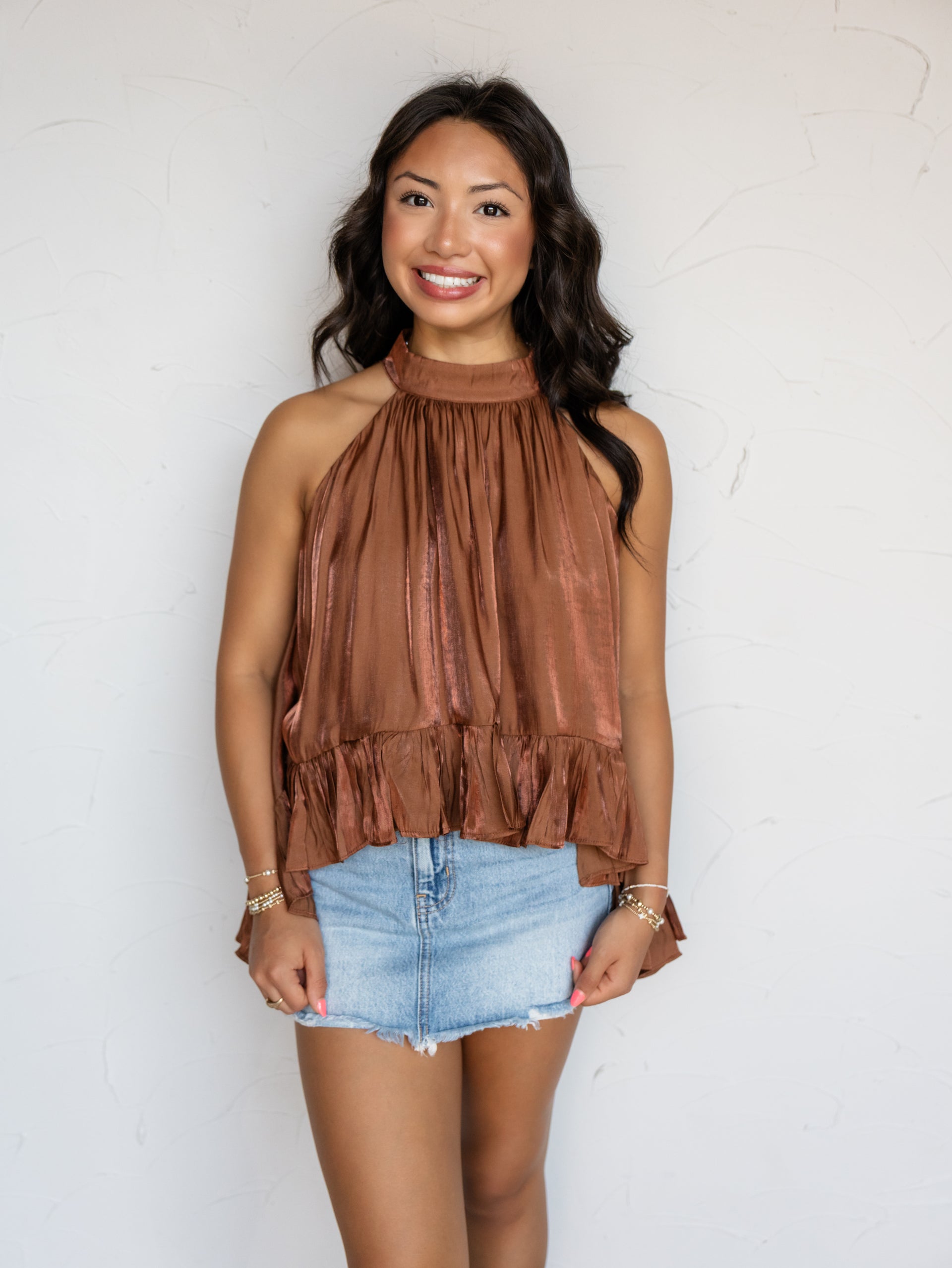 A three-quarter length photograph of a smiling woman with long wavy dark brown hair posing against a textured white wall. She is wearing a sleeveless bronze satin halter top with gathered pleating and a ruffled peplum hem, made from a 70% rayon and 30% polyester blend fabric, paired with a light-wash denim skirt and styled with stacked gold bracelets.