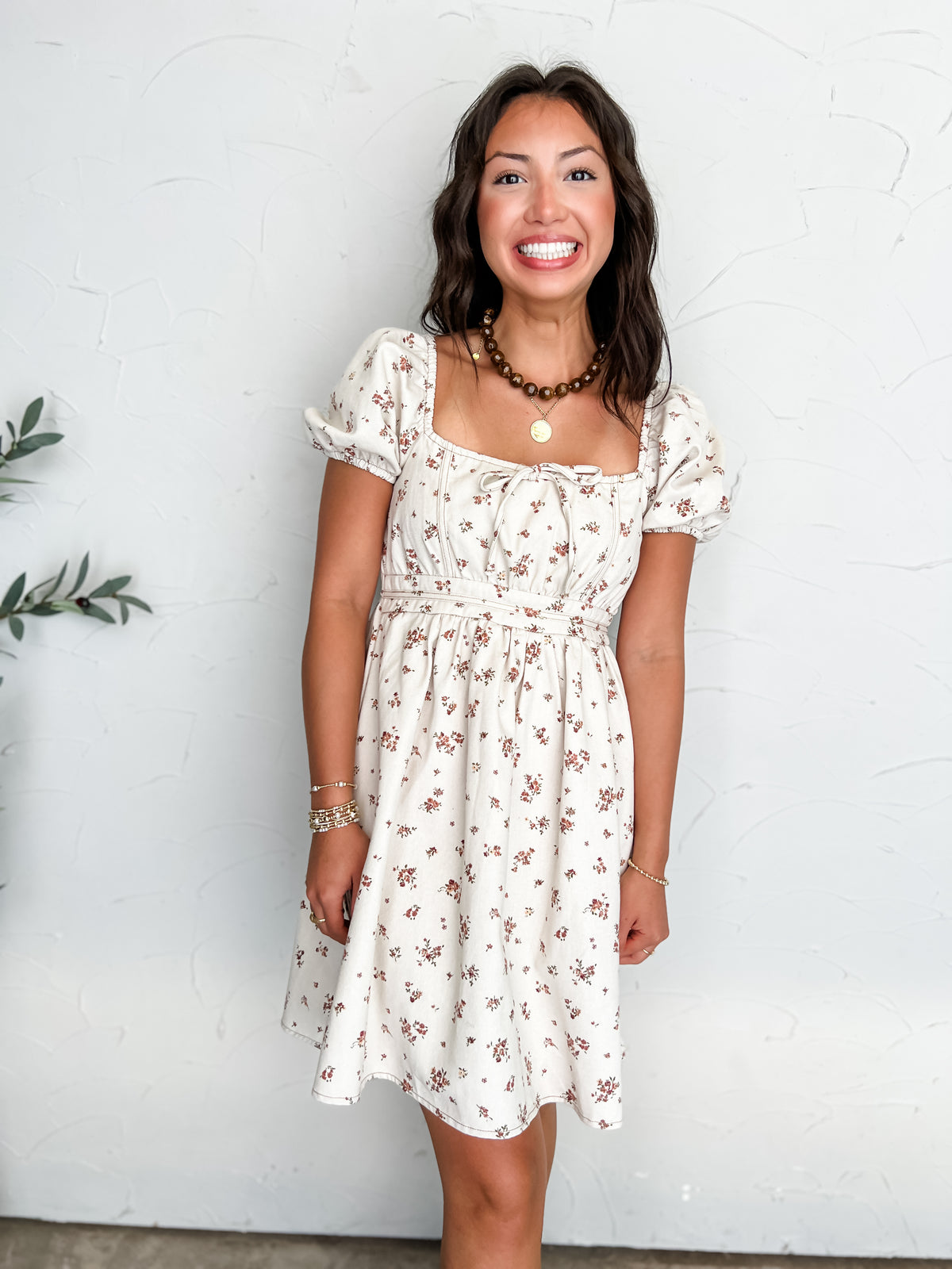 A portrait photograph of a smiling brunette woman with long wavy hair posing against a textured off-white wall next to a potted olive plant. She is wearing a short-sleeved mini dress made from 100% cotton in an off-white color with a delicate rust brown floral print, featuring a square neckline with a small front bow accent, puff sleeves, and a defined waistline, styled with a brown beaded choker, a gold coin pendant necklace, stacked beaded bracelets, and gold rings.