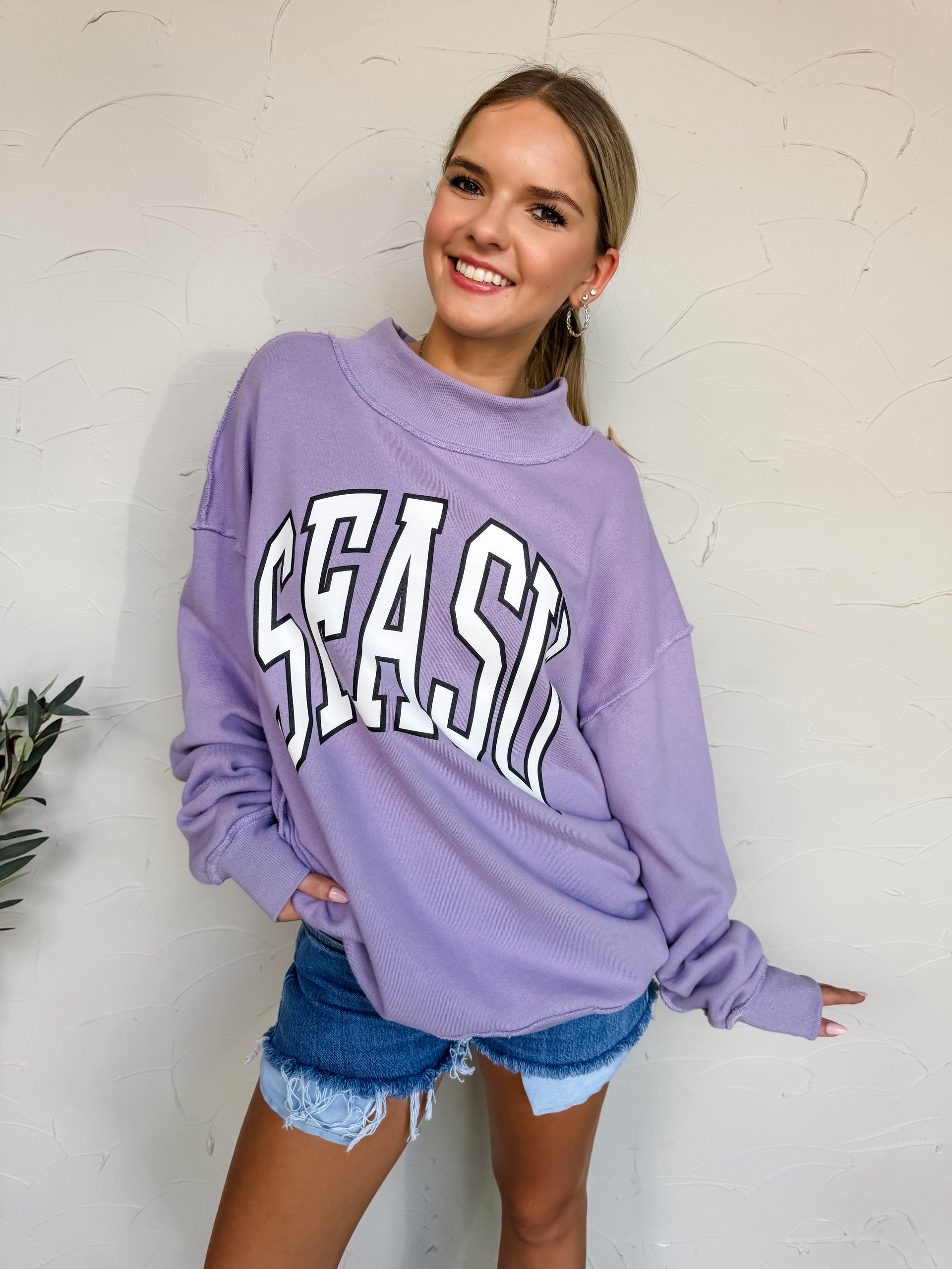 Woman wearing a purple sweatshirt with 'SEAS' text, standing against a white wall.