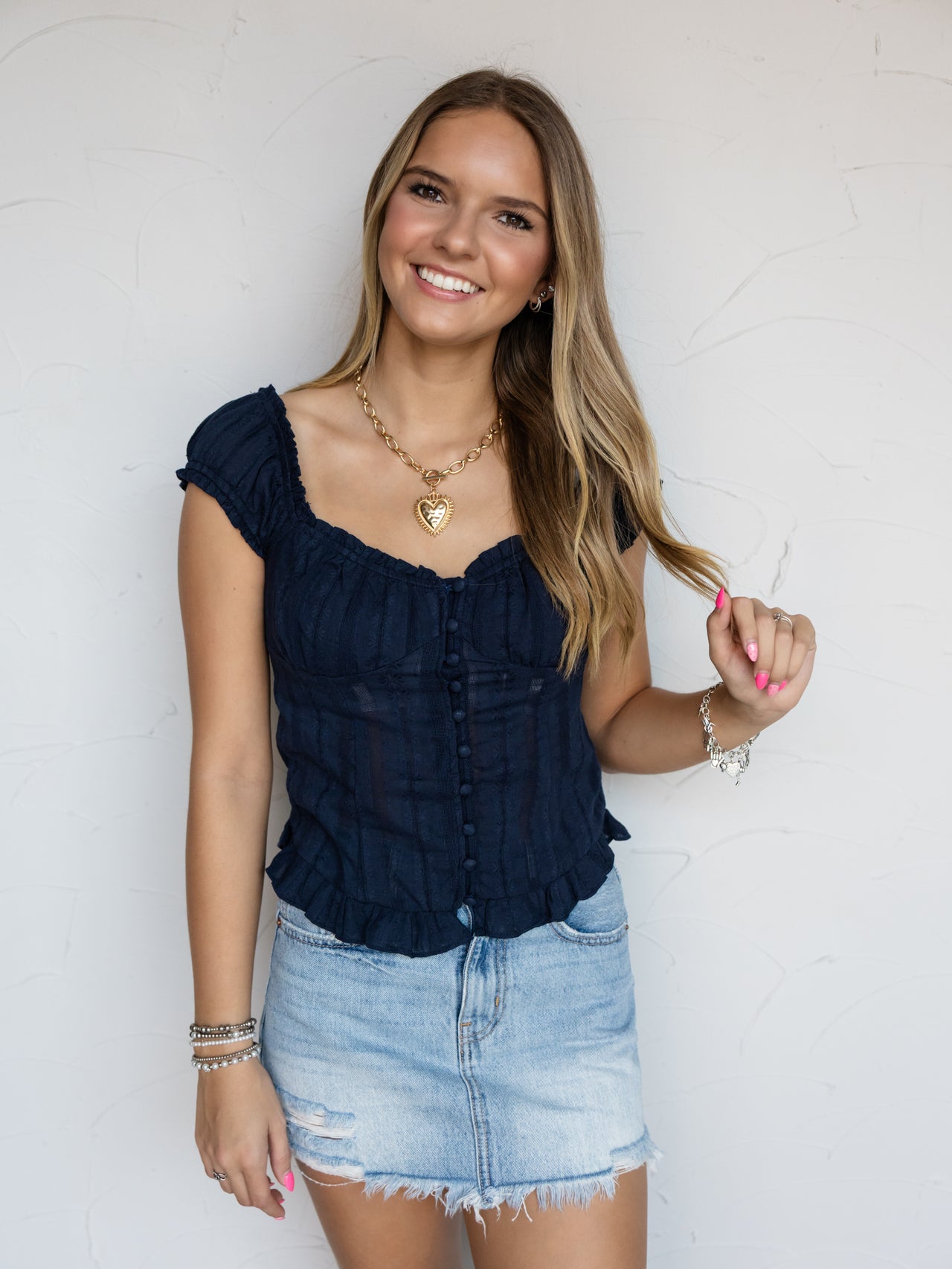 A three-quarter length photograph of a smiling woman with long blonde hair holding a strand of her hair while posing against a textured white wall. She is wearing a navy blue short-sleeved top made from 100% polyester fabric featuring a square neckline, decorative covered buttons down the center front, cap sleeves, and a small ruffled hem, paired with a light-wash distressed denim skirt and styled with a chunky gold heart necklace and bracelets.
