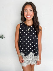 A portrait photograph of a smiling brunette woman posing against a textured off-white wall. She is wearing a two-piece matching set made from an 81% viscose and 19% nylon blend in a black and white polka dot print, featuring a sleeveless crewneck top with white eyelash lace trim along the hem, paired with matching polka dot shorts with white lace hemline detailing, styled with a brown beaded choker, a round gold pendant necklace, stacked gold bracelets, and gold rings.