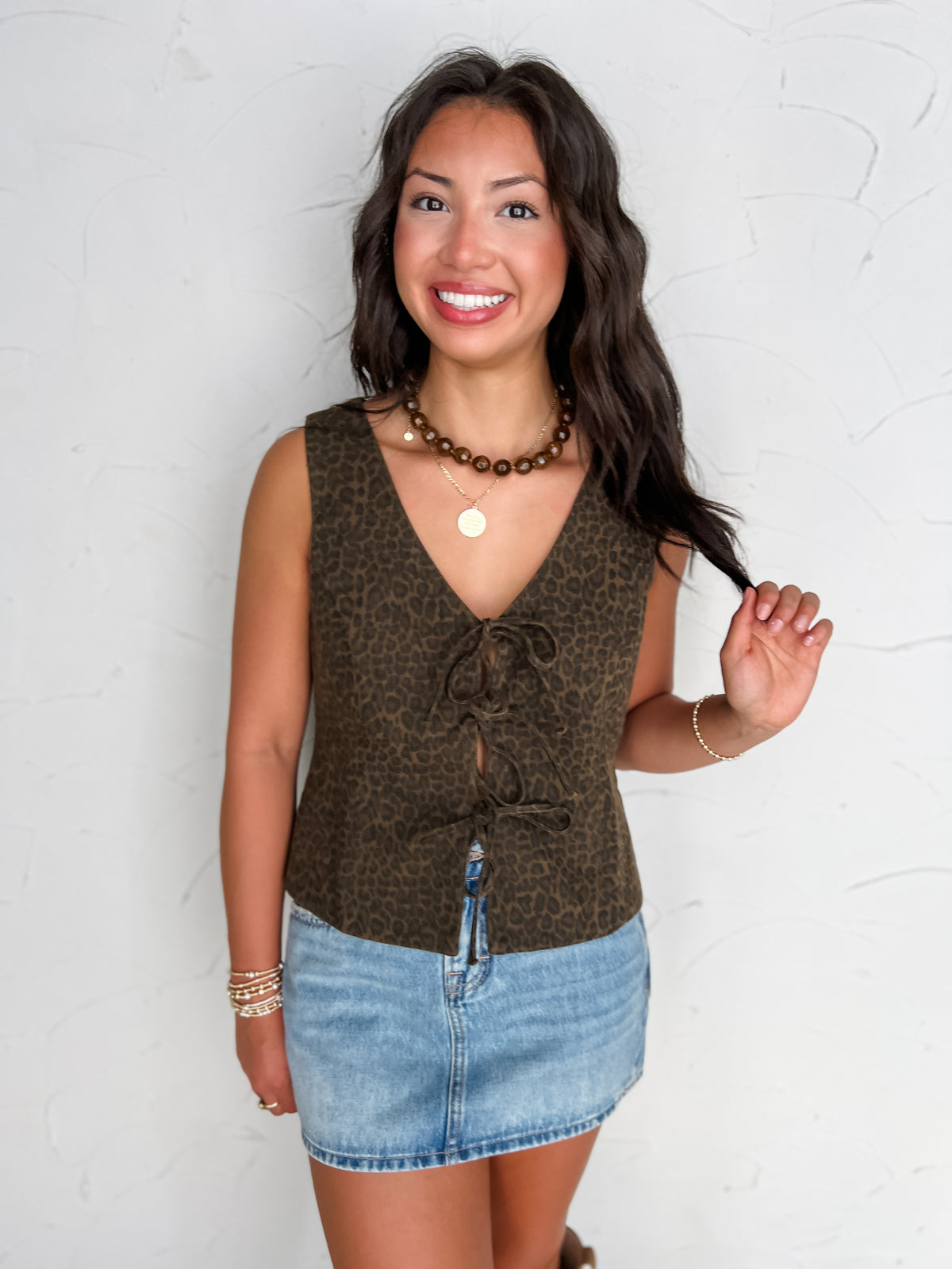 A portrait photograph of a smiling brunette woman posing while gently holding a lock of her hair against a textured off-white wall. She is wearing an olive green leopard print denim vest top made from 100% cotton, featuring a V-neckline, sleeveless cut, and three front fabric ties, paired with a light-wash denim skirt, styled with a brown beaded choker necklace, a gold coin pendant necklace, stacked gold bracelets, and gold rings.