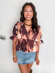A portrait photograph of a smiling brunette woman with long wavy hair posing against a textured off-white wall. She is wearing a short-sleeved surplice peplum top made from 100% cotton featuring an abstract brown, peach, and pink print with eyelet embroidery, ruffled shoulder trim, and ruffled cuffs, paired with a light-wash denim skirt, styled with a brown beaded choker necklace, a gold pendant chain, stacked gold bracelets, and rings.