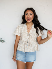 A three-quarter length photograph of a smiling woman with long dark wavy hair holding a strand of her hair while posing against a textured white wall. She is wearing a beige short-sleeved blouse made from 100% polyester fabric featuring white floral embroidery, scalloped edges, puff sleeves, double tie-front closures, and a peplum hem, paired with light-wash denim shorts and styled with a pearl choker necklace and stacked bracelets.