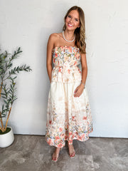 A full-length photograph of a smiling woman with long highlighted hair posing against a textured white wall next to a potted olive plant. She is wearing a two-piece matching set made from 100% cotton in a cream floral print with coral pink and soft blue flowers, consisting of a strapless smocked peplum top and a high-waisted midi skirt with side pockets, paired with clear strap heels, styled with a chunky white pearl necklace, a dainty gold cross chain, and silver bracelets.
