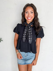 A portrait photograph of a smiling brunette woman posing against a textured off-white wall. She is wearing a black short-sleeved top made from 100% polyester, featuring a button-down front, pointed collar, puff sleeves, and a peplum hem, paired with a light wash denim skirt, styled with a black and white polka dot scarf tied at her neck, a brown beaded necklace, and stacked beaded bracelets.