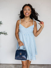 A three-quarter length photograph of a smiling woman with long dark wavy hair holding a strand of her hair while posing against a textured white wall. She is wearing a light blue and white vertical striped cami mini dress made from 100% polyester fabric, featuring a V-neckline, thin shoulder straps, an empire waist, and a horizontal-striped ruffled hem, styled with a glossy navy croc-embossed handbag, a pearl choker necklace, and stacked gold bracelets.