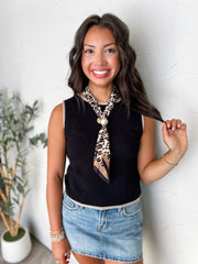 A portrait photograph of a smiling brunette woman holding a strand of her hair while posing against a textured off-white wall next to a potted olive plant. She is wearing a sleeveless black knit tank top made from a 70% viscose and 30% polyester blend, featuring light beige contrast ribbing along the crew neck, armholes, and bottom hem, paired with a light-wash denim skirt, styled with a leopard print silk neck scarf secured with a large pearl slide, a delicate gold beaded bracelet, and stacked beaded brace